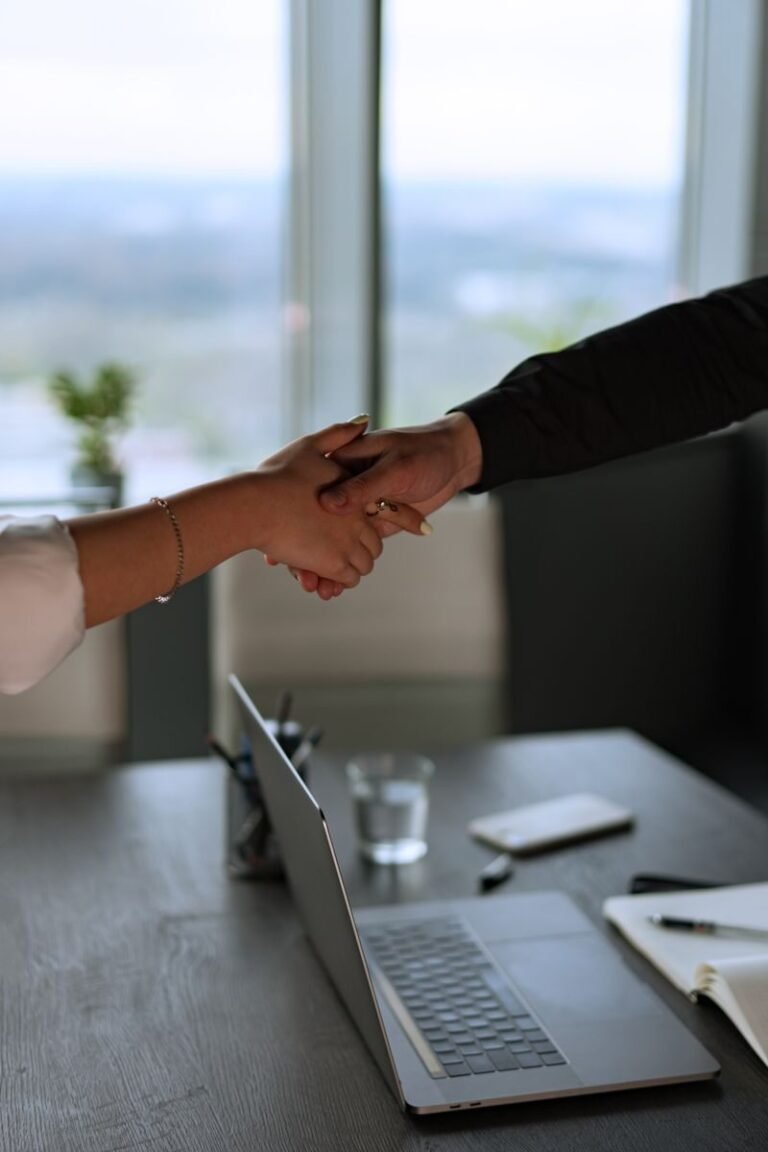 Two business professionals shaking hands across a desk with a laptop